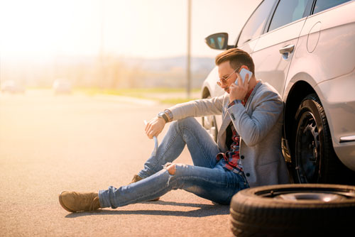 Junger Mann sitzt mit Radschlüssel neben seinem Auto und telefoniert